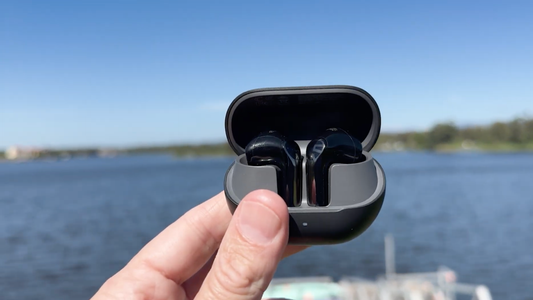 A hand holding an open charging case featuring the black Soundpeats Air6 HS wireless earbuds, set against a blurred background of a blue lake and clear sky.