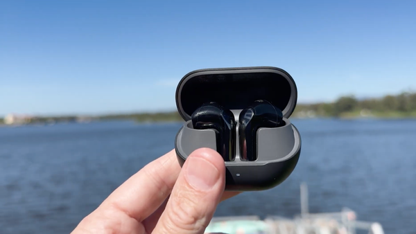 A hand holding an open charging case featuring the black Soundpeats Air6 HS wireless earbuds, set against a blurred background of a blue lake and clear sky.