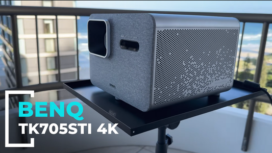 A side-angle shot of a grey BenQ TK705STi projector resting on a flat tray mount. The background shows a balcony railing with a blurred view of a coastal building and the ocean.